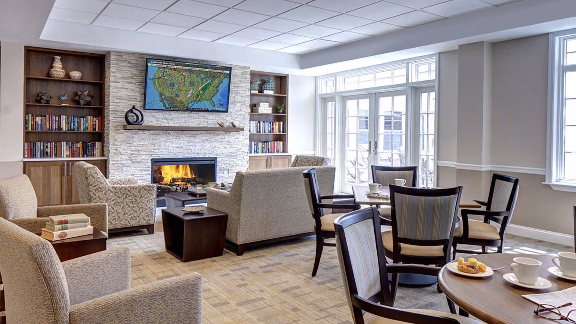 Comfortable senior living common area with sofas and chairs arranged around a fireplace and TV, bookshelves, and tables by large windows.