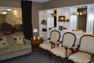 Communal living room with a sofa, three upholstered armchairs, a side table with a lamp, and built-in shelving displaying decorative items.