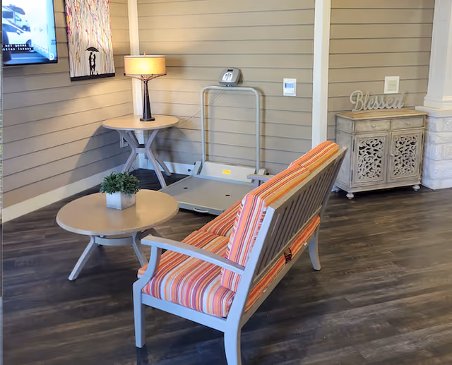 A cozy seating area with a striped cushioned bench facing a round table with a small potted plant. Behind the bench is a side table with a lamp and a wall-mounted TV. To the right, there is a decorative cabinet with the word 'Blessed' on top. The floor is dark wood, and the walls have horizontal paneling.