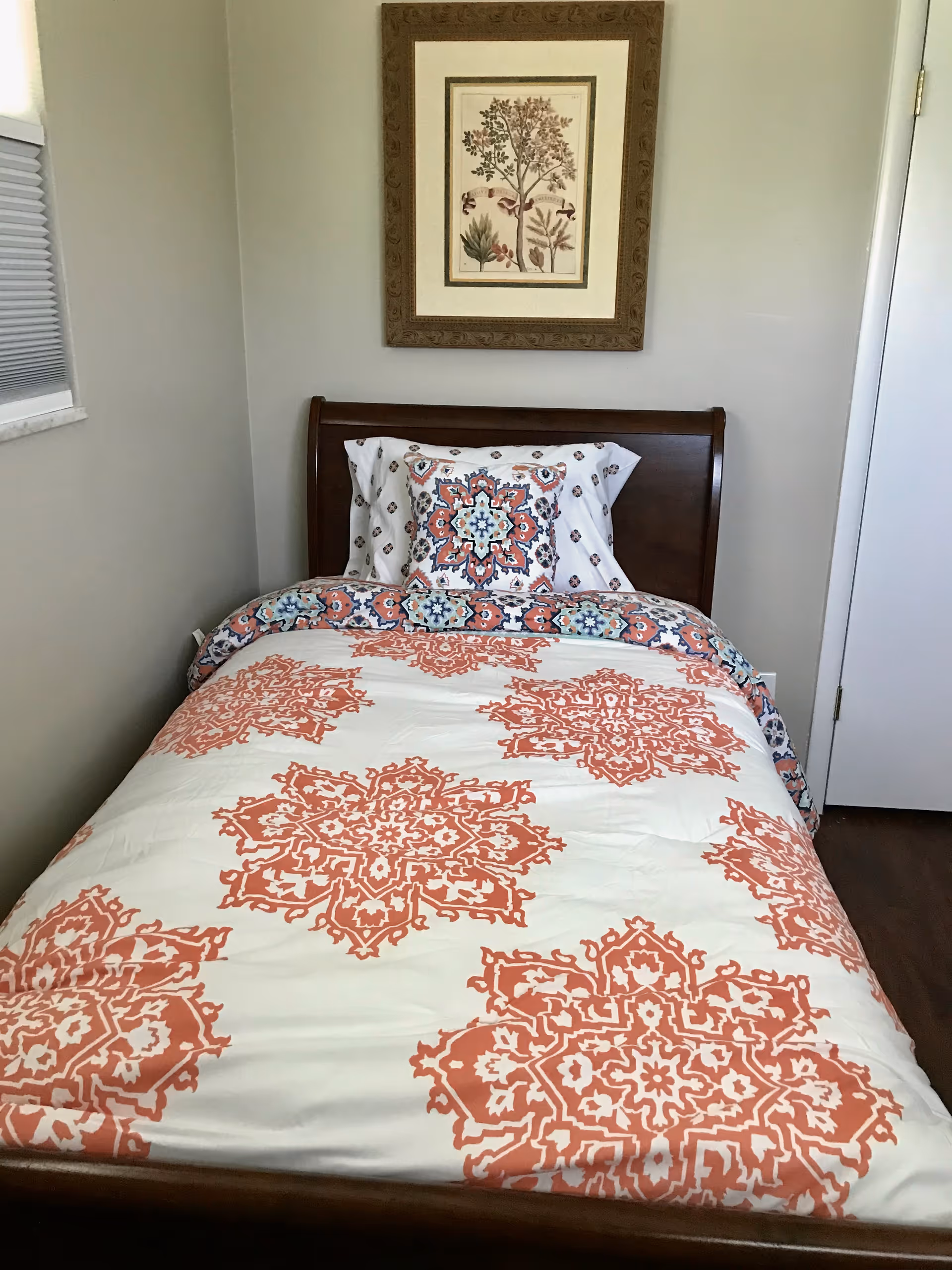 A single bed with a wooden headboard, covered with a white and orange patterned comforter and matching pillow. Above the bed hangs a framed botanical print. To the left is a window with closed blinds, and to the right is a closed white door.