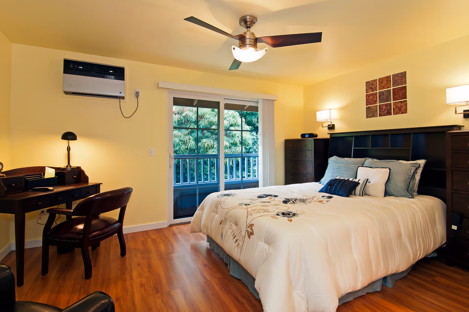 A cozy bedroom with a large bed featuring a white comforter with floral patterns and multiple pillows. The room has wooden flooring, a ceiling fan with light, a small desk with a chair and lamp, and a dresser. There is a sliding glass door leading to a balcony with greenery outside. The walls are painted yellow and decorated with a small wall art piece above the bed.