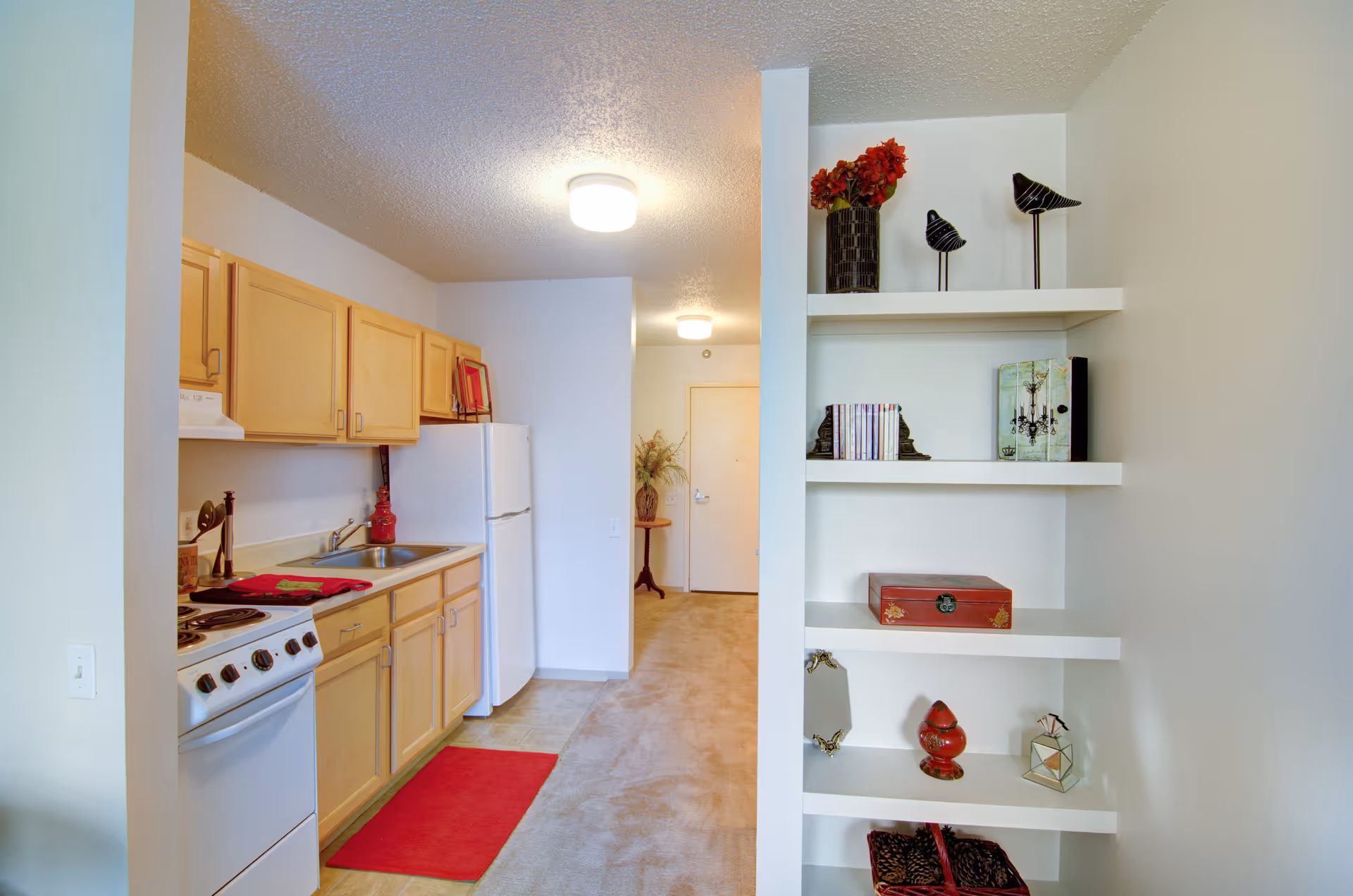 Interior view of a senior living facility kitchen area with light wood cabinets, a white stove, and a white refrigerator. To the right, there is a built-in white shelving unit decorated with books, vases, and small decorative items. The floor transitions from tile in the kitchen to carpet in the hallway leading to a closed door.