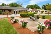 Outdoor courtyard area with a concrete patio featuring a metal table and chairs surrounded by flower beds with blooming pink and white flowers. In the background, there are single-story buildings with large windows and a gazebo, under a partly cloudy blue sky.