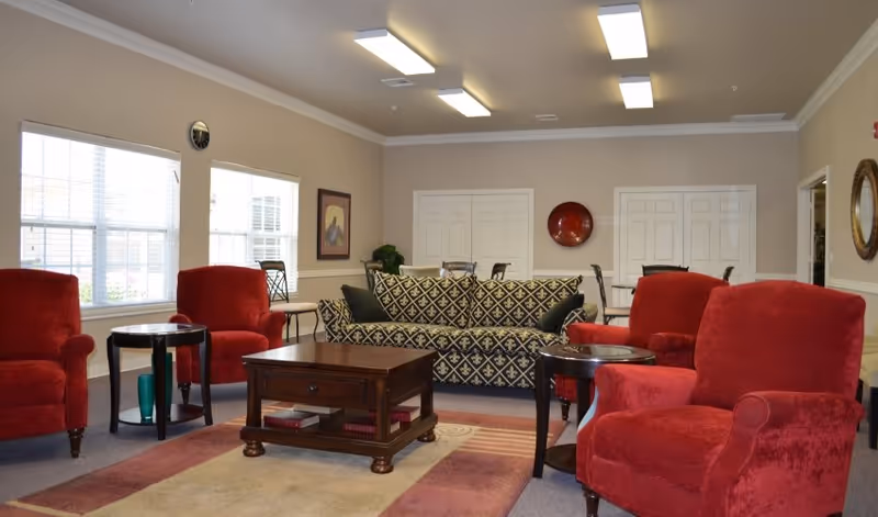 Spacious communal living room with red upholstered armchairs, a patterned sofa, wooden coffee table and large windows.