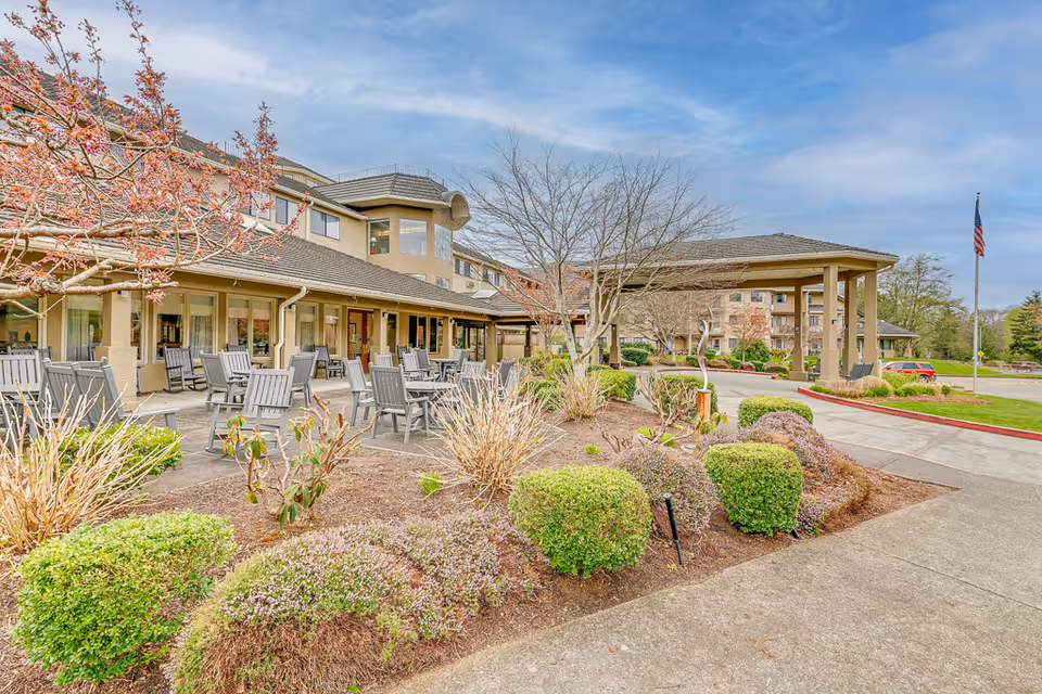 Front exterior of a senior living facility showing the entrance with a covered porte-cochere, outdoor seating area, and landscaped grounds.