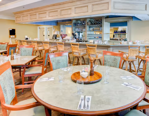 Interior view of a dining area at Franke At Seaside featuring a round table set with glasses, napkins, and cutlery. Surrounding the table are cushioned chairs with wooden frames. In the background, there is a bar counter with high stools, shelves stocked with bottles and glassware, and a mirrored wall.