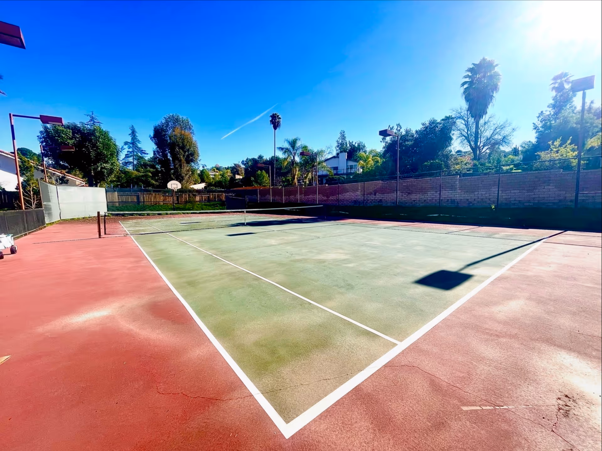 Empty outdoor tennis court with a net, basketball hoop, and palm trees under a clear blue sky.