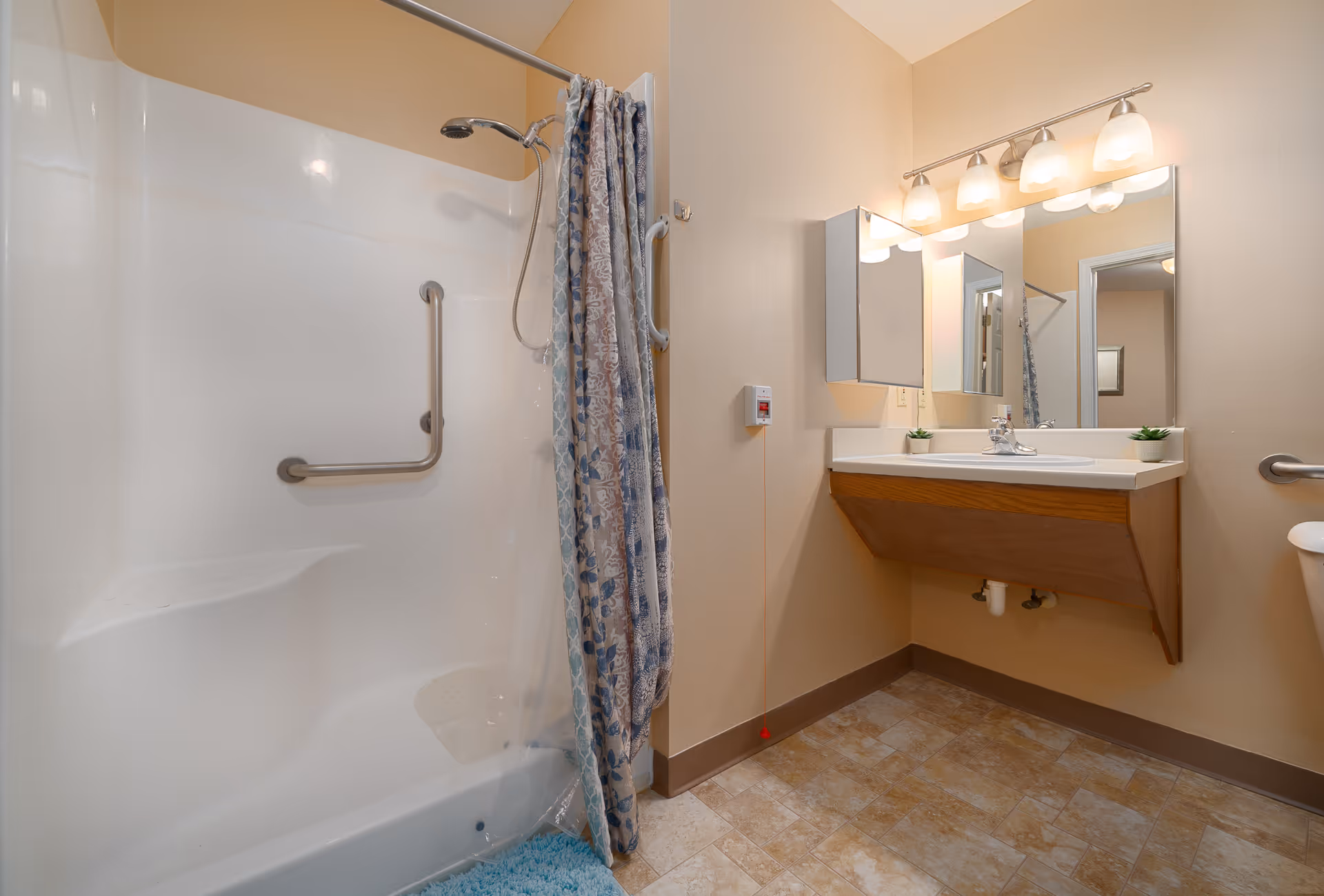 A bathroom with a white bathtub and shower combination featuring a floral patterned shower curtain. The bathroom has beige walls, a vanity with a sink, a large mirror, and four light fixtures above the mirror. There are grab bars installed near the bathtub and next to the toilet for accessibility. The floor is covered with beige and brown tiles.