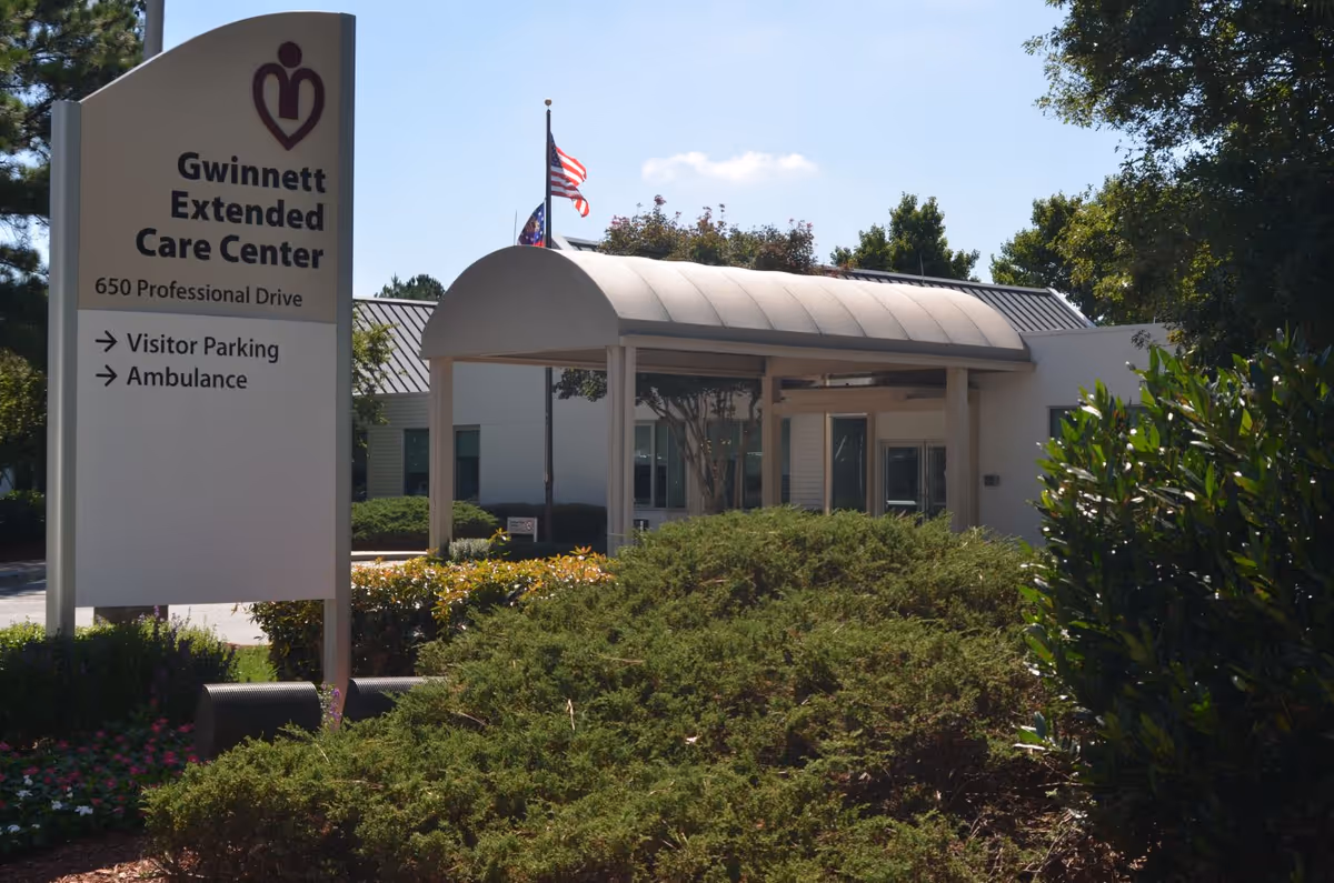 Exterior view of Gwinnett Extended Care Center showing the entrance with a covered walkway, surrounded by greenery and bushes. A sign in the foreground displays the facility name, address, and directions for visitor parking and ambulance. An American flag is visible in the background.
