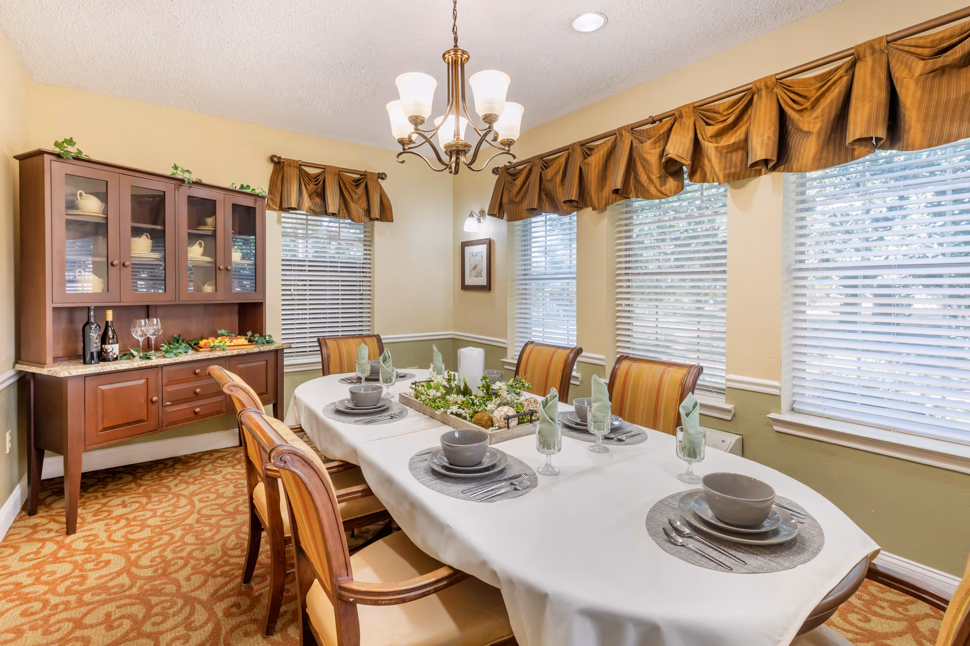 A dining room with a long table covered with a white tablecloth, set with gray dishes, bowls, glasses with folded green napkins, and silverware. The table has a centerpiece with candles and decorative greenery. There are six wooden chairs with striped cushions around the table. A wooden hutch with glass doors displaying dishes and wine bottles is against the wall. The room has beige walls, three windows with white blinds and brown valances, and a chandelier with five lights hanging from the ceiling.