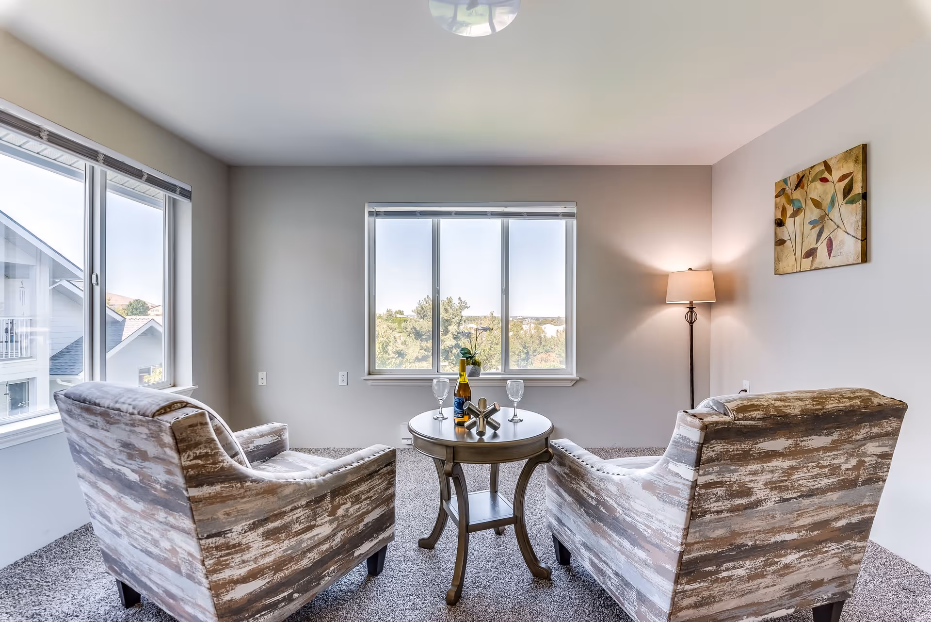 A bright living room with two patterned armchairs facing a round wooden table with two wine glasses and a bottle. The room has large windows showing an outdoor view, a floor lamp in the corner, and a colorful leaf-themed painting on the wall.