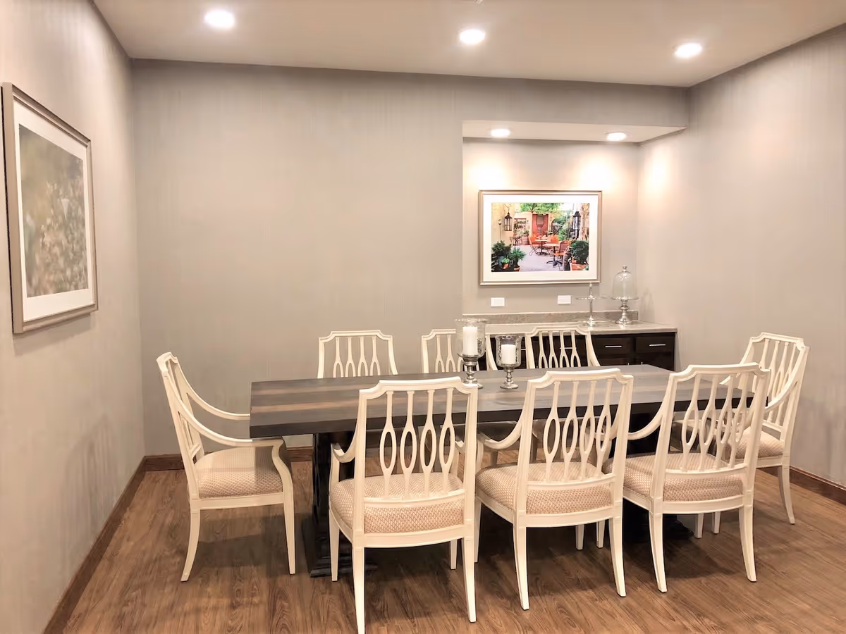 A dining room with a long rectangular wooden table surrounded by eight white cushioned chairs. The room has light gray walls, wooden flooring, and recessed ceiling lights. There is a framed picture on the back wall above a small sideboard with decorative items, and another framed artwork on the left wall.