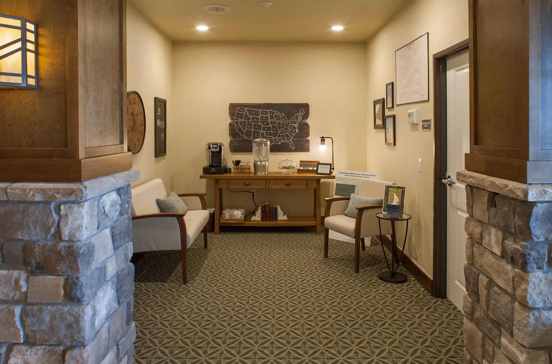 A cozy seating area in a memory care facility with two cushioned chairs facing each other, a wooden table against the back wall holding a coffee maker, water dispenser, and other small items. The walls are decorated with framed pictures and a wooden map of the United States. The floor is carpeted with a geometric pattern, and stone pillars frame the entrance to the area.