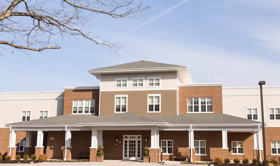 Front exterior view of a multi-story senior living facility building with a covered entrance, brick and siding facade, multiple windows, and a clear sky with tree branches visible.