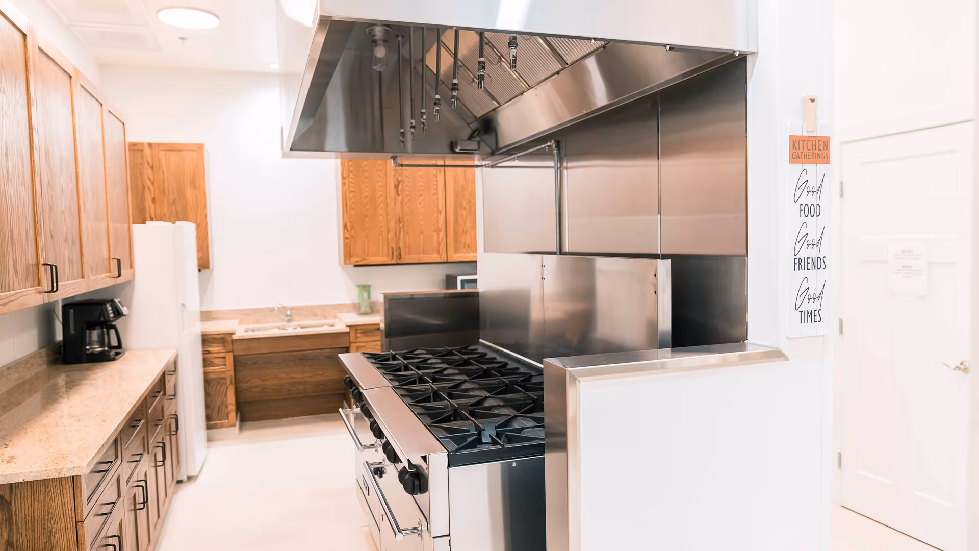 Commercial-style kitchen with a large stainless-steel stove and hood, wooden cabinets, countertops, a refrigerator, and a coffee maker.