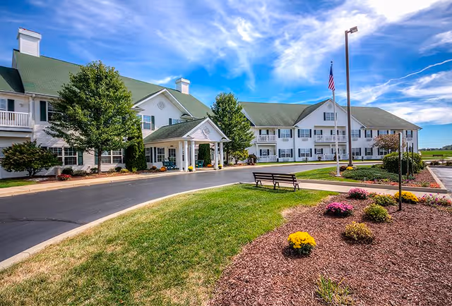 Exterior view of Jefferson Manor, a large white building with green roofs, surrounded by landscaped gardens, trees, and a paved driveway. There is a bench on the grass near a flower bed with colorful flowers, and an American flag on a flagpole near the entrance. The sky is blue with some clouds.