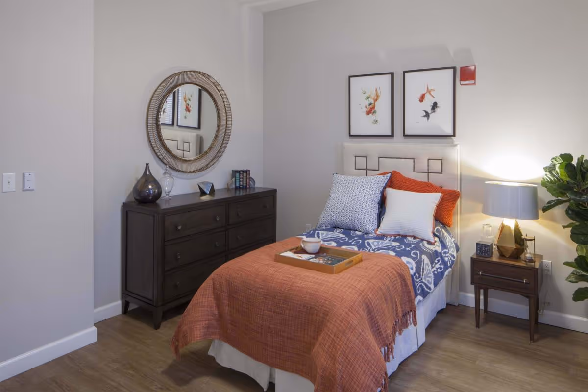 A neatly staged single bedroom featuring a bed with an orange throw and decorative pillows, a dresser with a round mirror, and a nightstand with a lamp.