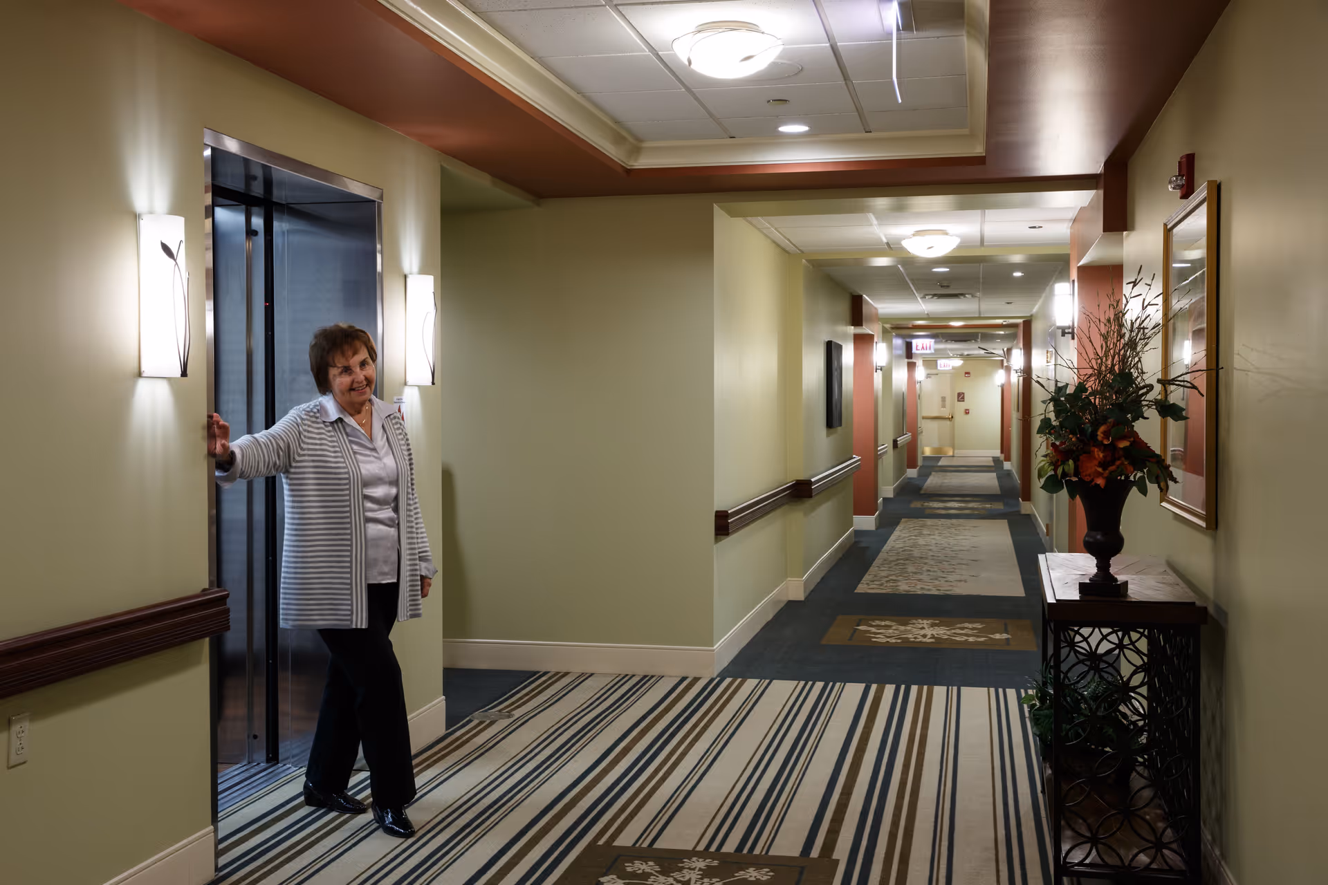 An elderly woman stands at the open door of an elevator in a well-lit hallway of a senior living facility. The hallway features patterned carpeting, light green walls with wooden handrails, wall sconces, and a decorative table with a flower arrangement. The ceiling has recessed lighting and a drop ceiling design.