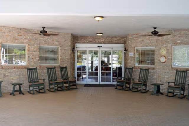 Covered entrance area with brick walls featuring two windows and a set of automatic sliding glass doors in the center. There are several green rocking chairs and small tables arranged along the walls. Two ceiling fans and three ceiling lights are mounted on the white ceiling.
