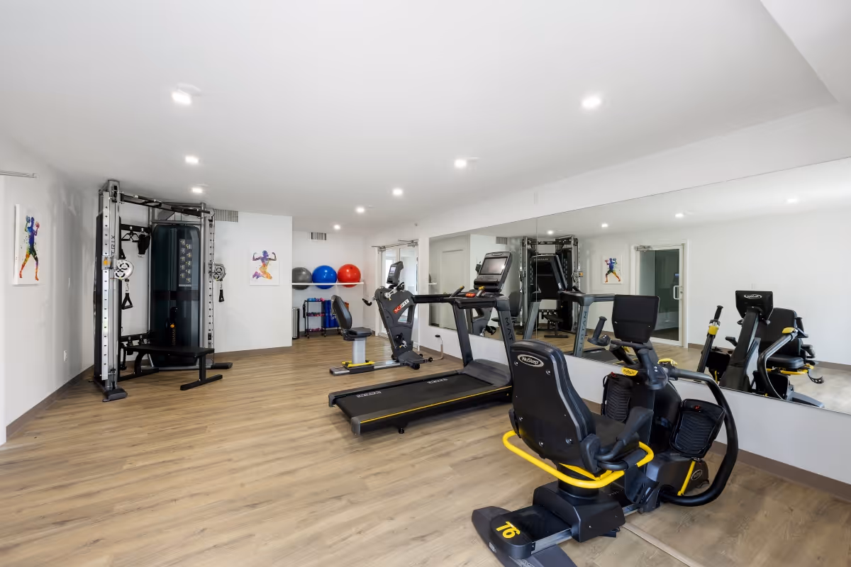 A bright and spacious fitness room with exercise equipment including a treadmill, recumbent bike, and a multi-functional weight machine. The room has wooden flooring, white walls, recessed ceiling lights, and a large mirror covering one wall. Colorful exercise balls and fitness accessories are visible in the background.