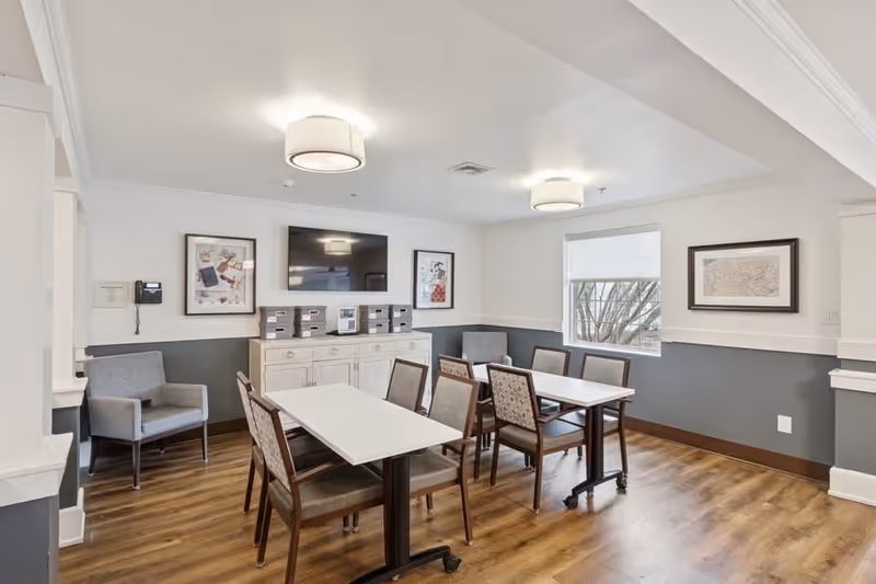 A bright and clean dining room with two rectangular white tables and eight chairs arranged around them. The room has wood flooring, gray and white walls, and a large window letting in natural light. There are two gray armchairs in the corners, a white sideboard with storage boxes, a wall-mounted flat-screen TV, and framed artwork on the walls.