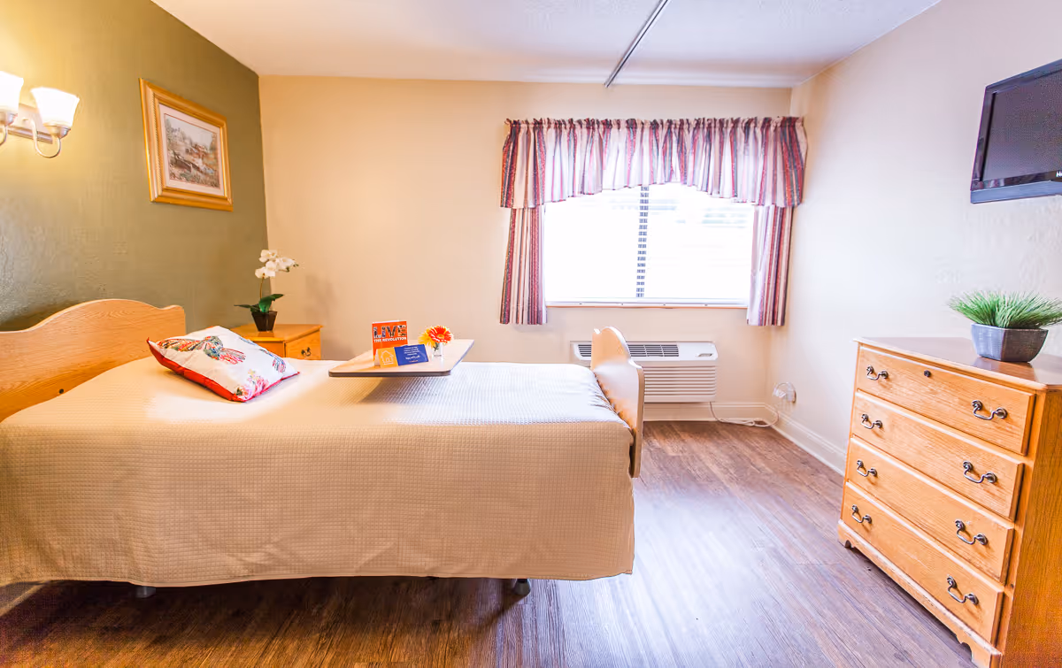 A tidy single bedroom with a bed, bedside table, wooden dresser, window with curtains, and a wall-mounted TV.