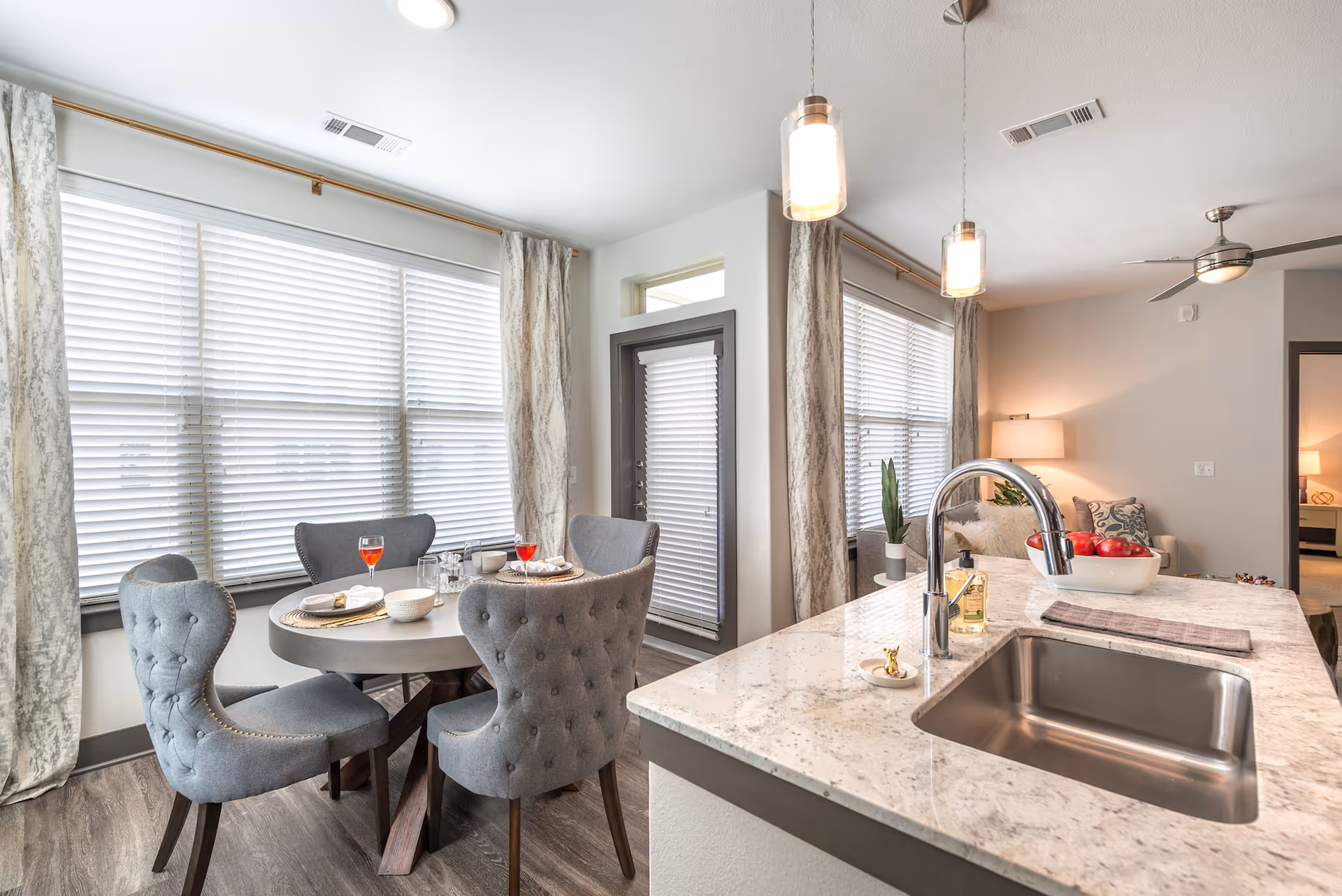 Bright and modern dining area with a round table set for four, featuring gray upholstered chairs. Adjacent to the dining area is a kitchen island with a marble countertop, a stainless steel sink, a chrome faucet, a bowl of red apples, and a soap dispenser. Large windows with white blinds and patterned curtains allow natural light to fill the space. In the background, a cozy living area with a lamp and ceiling fan is visible.