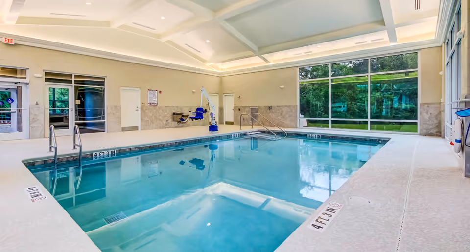 Indoor swimming pool with large windows, ceiling lighting, and an accessible pool lift in a senior living facility.