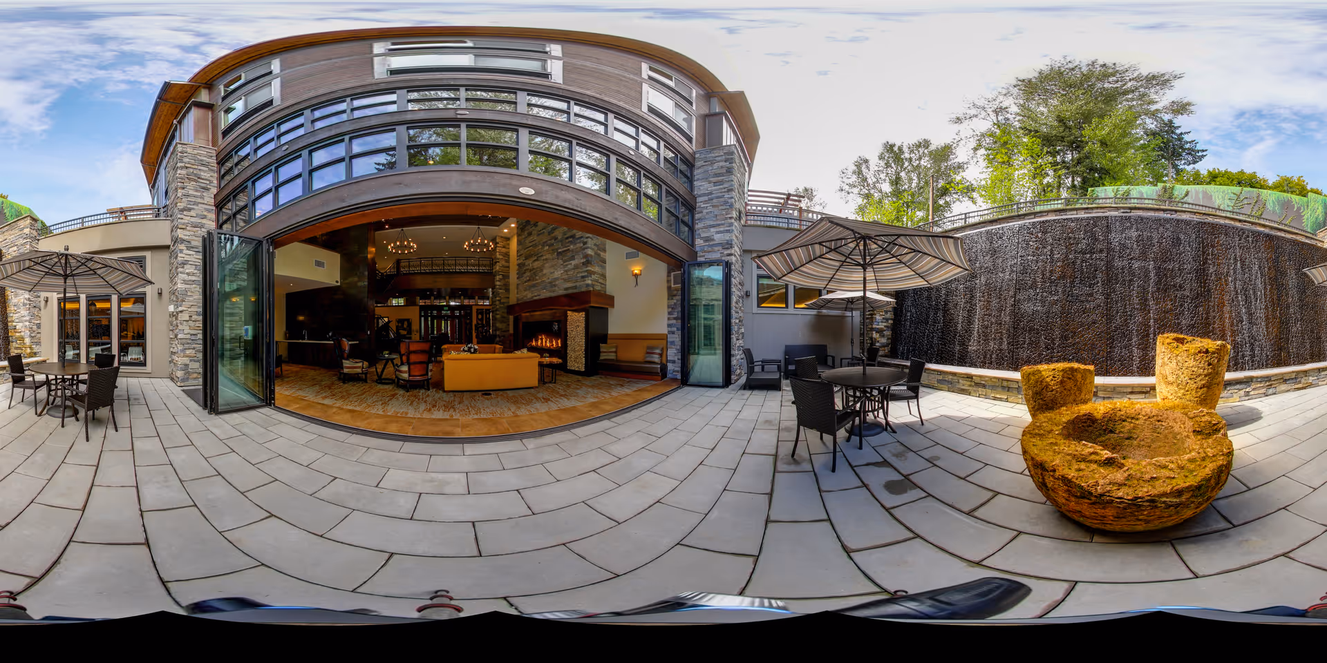 Outdoor patio area of Aegis Living Mercer Island featuring stone flooring, several tables with umbrellas, a large stone water fountain, and an open view into a cozy indoor lounge area with a fireplace and seating.