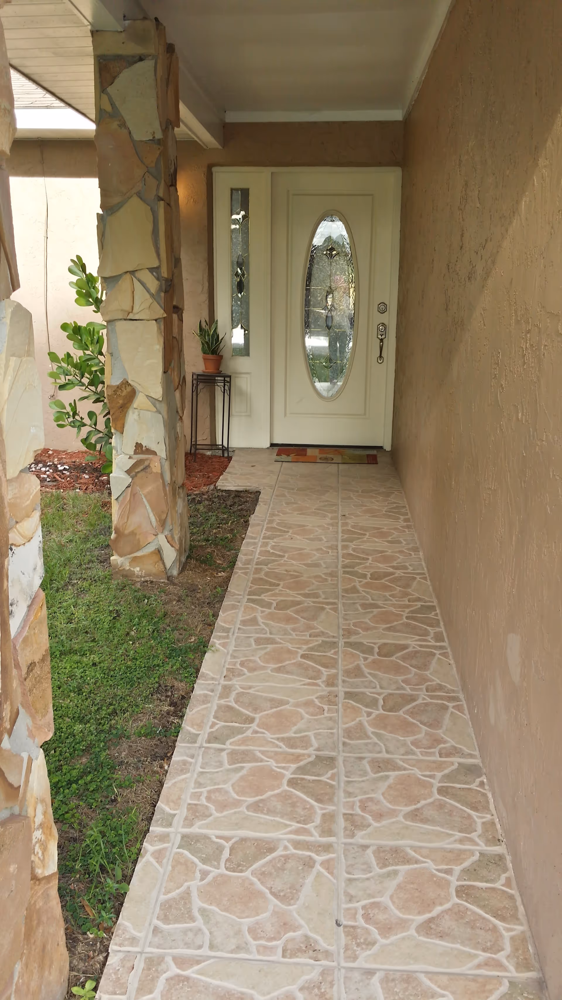 A tiled walkway leading to a white front door with decorative glass panels. The walkway is bordered by a stone pillar on the left and a beige textured wall on the right. There is a small plant on a stand near the door and some greenery visible to the left of the walkway.