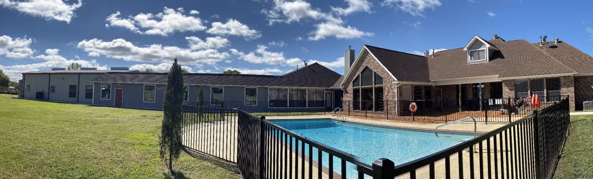 Panoramic view of a residential care facility exterior with a fenced outdoor swimming pool, brick building, and grassy lawn under a partly cloudy sky.