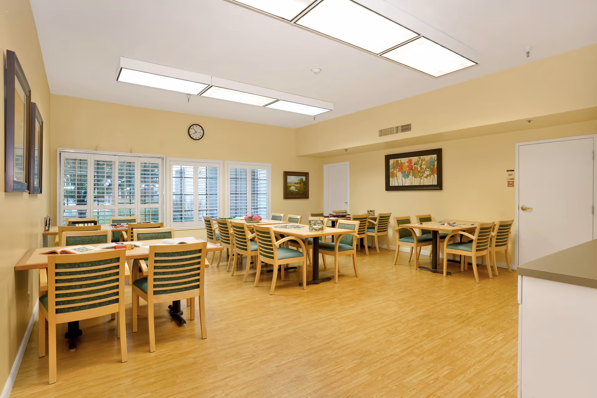 A bright dining room with several wooden tables and chairs arranged neatly. The room has light yellow walls, large windows with white shutters, and framed artwork on the walls. The floor is a light wood laminate, and there are ceiling lights providing ample illumination.