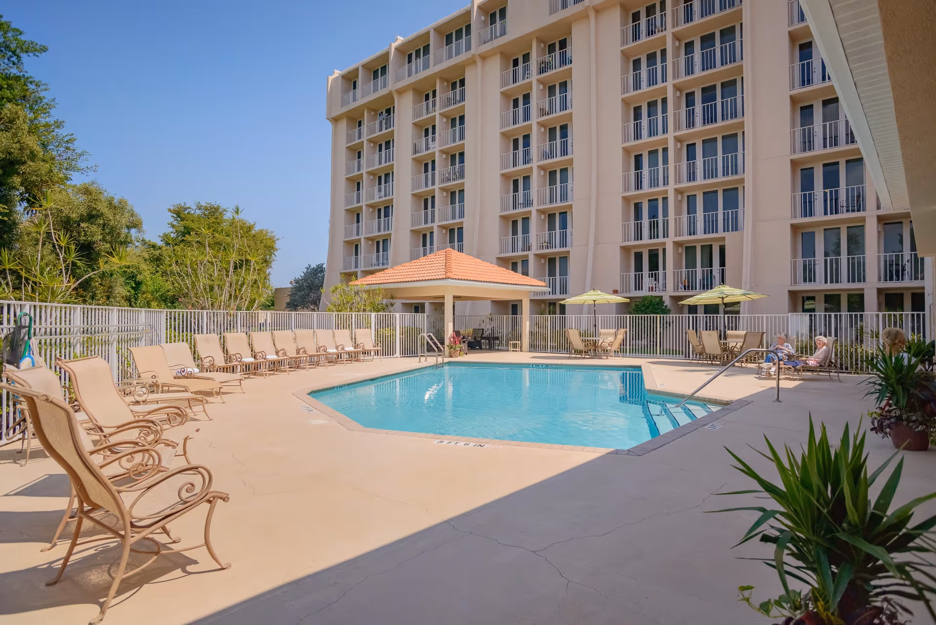 Outdoor swimming pool area at Garden Plaza at Inverrary with lounge chairs lined up along the poolside, a small covered pavilion with a tiled roof, tables with umbrellas, and a multi-story building in the background under a clear blue sky.