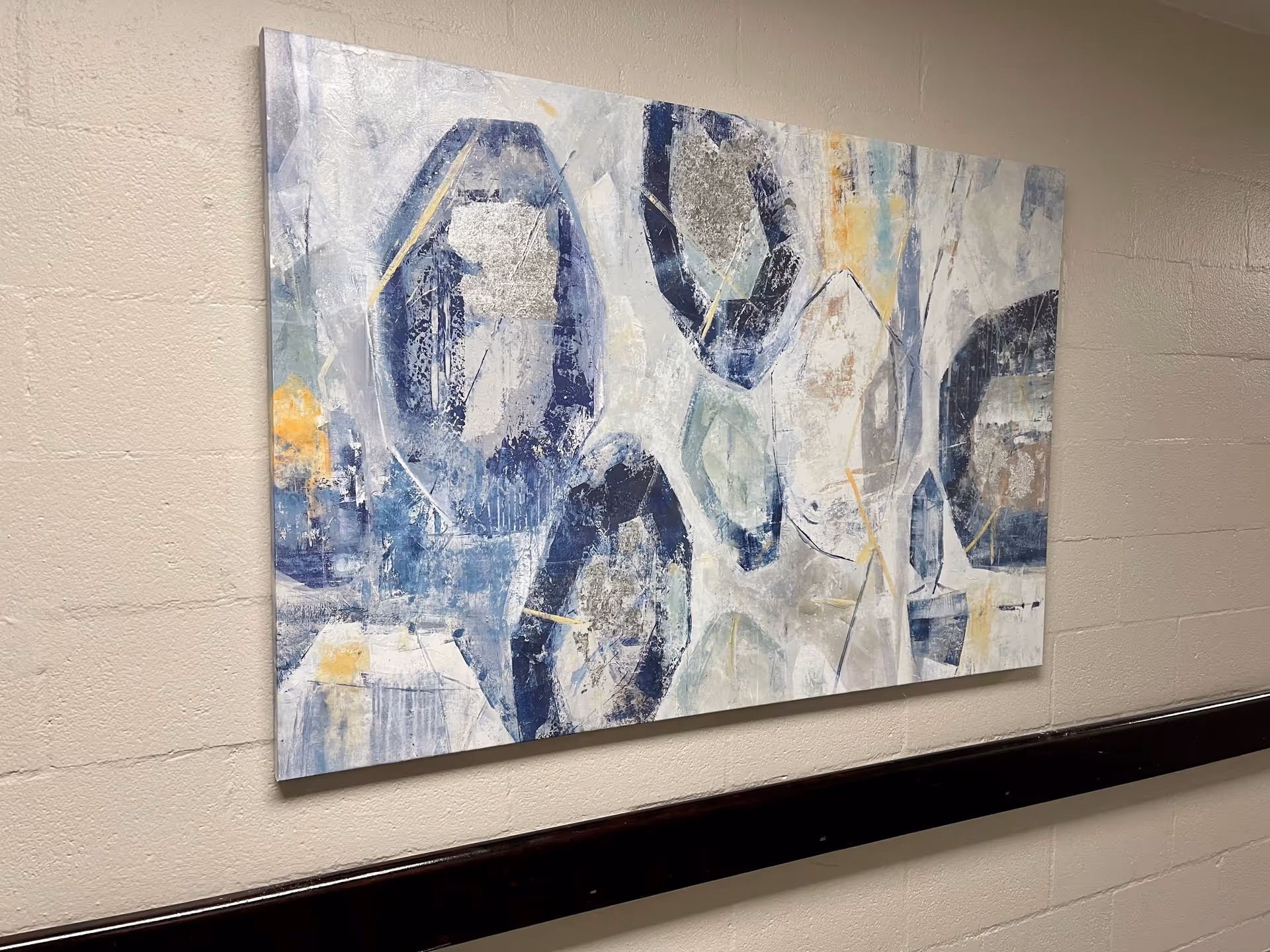 Abstract painting with blue, white, gray, and yellow tones hanging on a beige wall above a dark wooden handrail in a hallway.
