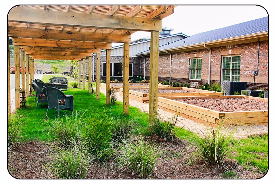 Outdoor garden area at Thomas Kelly Senior Living Community featuring a wooden pergola with green wicker chairs underneath, green grass, raised garden beds filled with soil, and a brick building in the background.