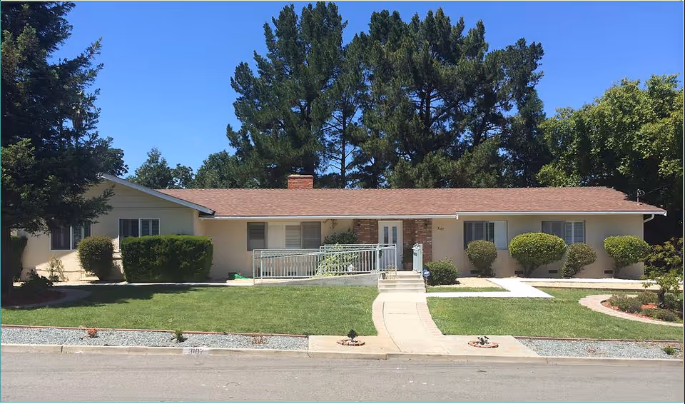 Single-story residential building with a brown roof and beige exterior walls, surrounded by green grass, trimmed bushes, and tall trees in the background under a clear blue sky. A concrete walkway leads to the front door, which has a small ramp and steps.