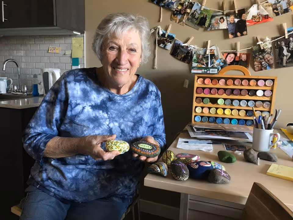 An elderly woman with short gray hair wearing a blue tie-dye shirt is sitting at a table in a room. She is smiling and holding two painted rocks. On the table in front of her are several more painted rocks, paintbrushes in a white mug, and a wooden paint palette with multiple colors. Behind her, photos are clipped to strings on the wall, and a kitchen sink and cabinets are visible in the background.