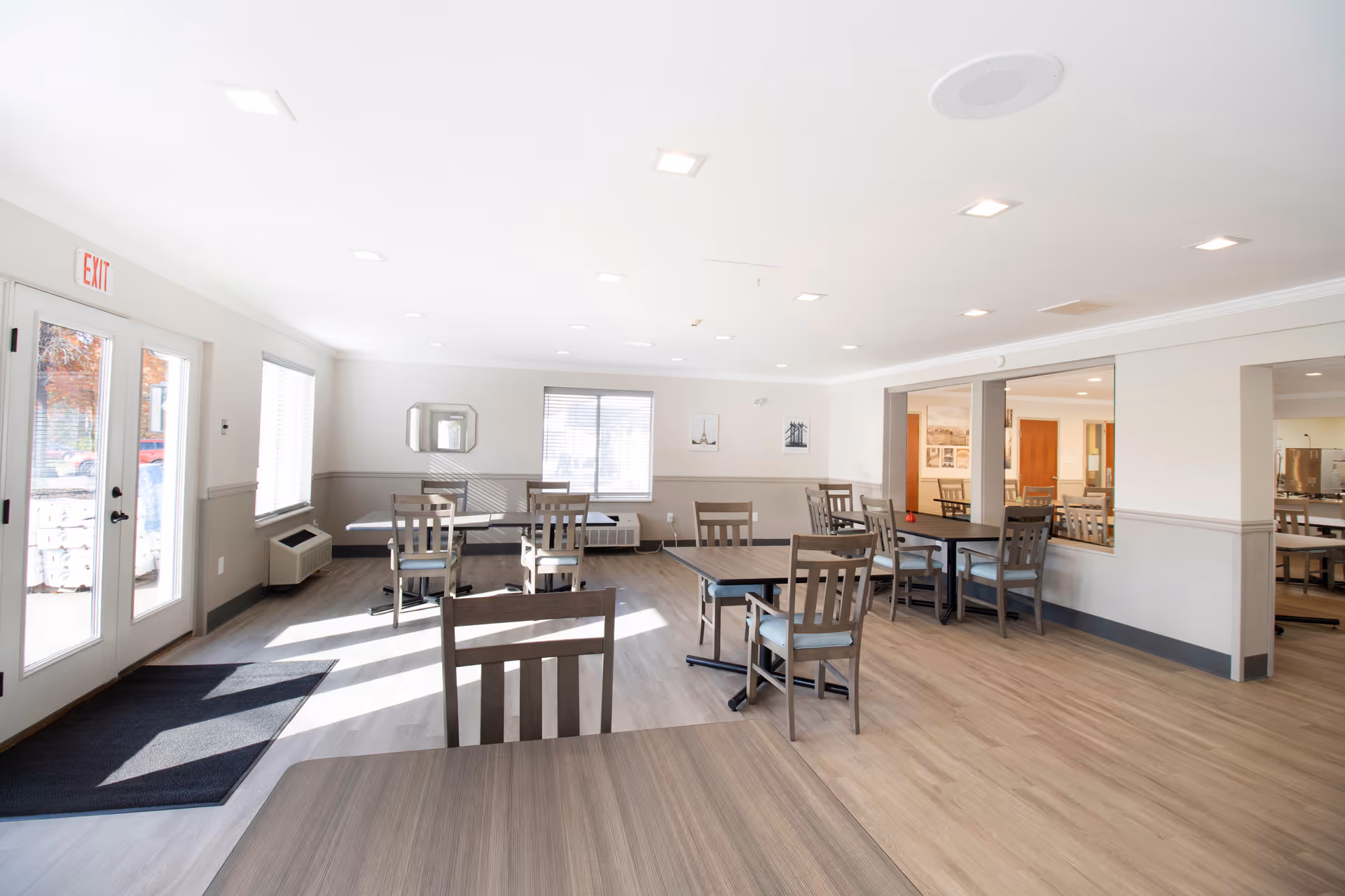 Bright and spacious dining room with multiple wooden tables and chairs arranged neatly. Large windows allow natural light to fill the room, and there is a glass door with an exit sign above it. The walls are painted white with a gray wainscoting, and the floor has light-colored wood laminate.