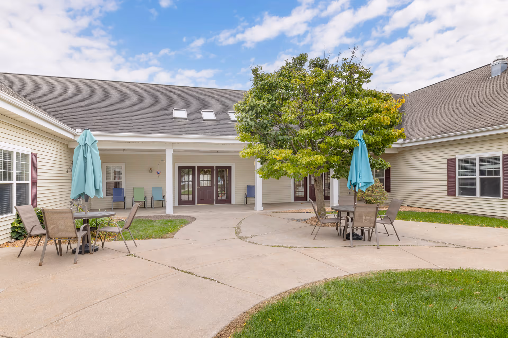 Outdoor courtyard area of Brookdale LaCrosse Assisted Living featuring a paved walkway, green grass, a tree in the center, and patio tables with chairs and closed turquoise umbrellas. The building exterior is beige with maroon doors and window shutters.