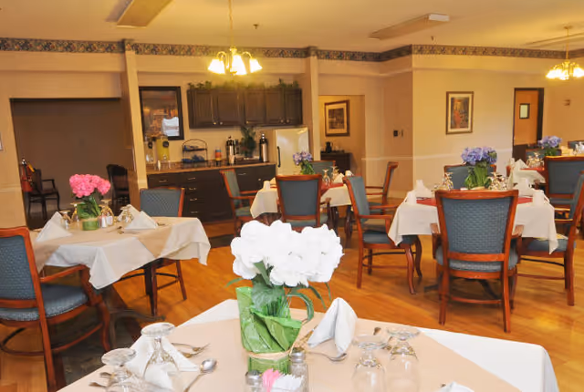 A dining room in a senior living facility with multiple tables covered in white tablecloths, each set with folded napkins, glasses, and silverware. Several tables have flower centerpieces in pink, white, and purple. The room has wooden floors, blue cushioned chairs with wooden frames, and a kitchenette area with dark cabinets and a white refrigerator in the background. Warm lighting fixtures hang from the ceiling.