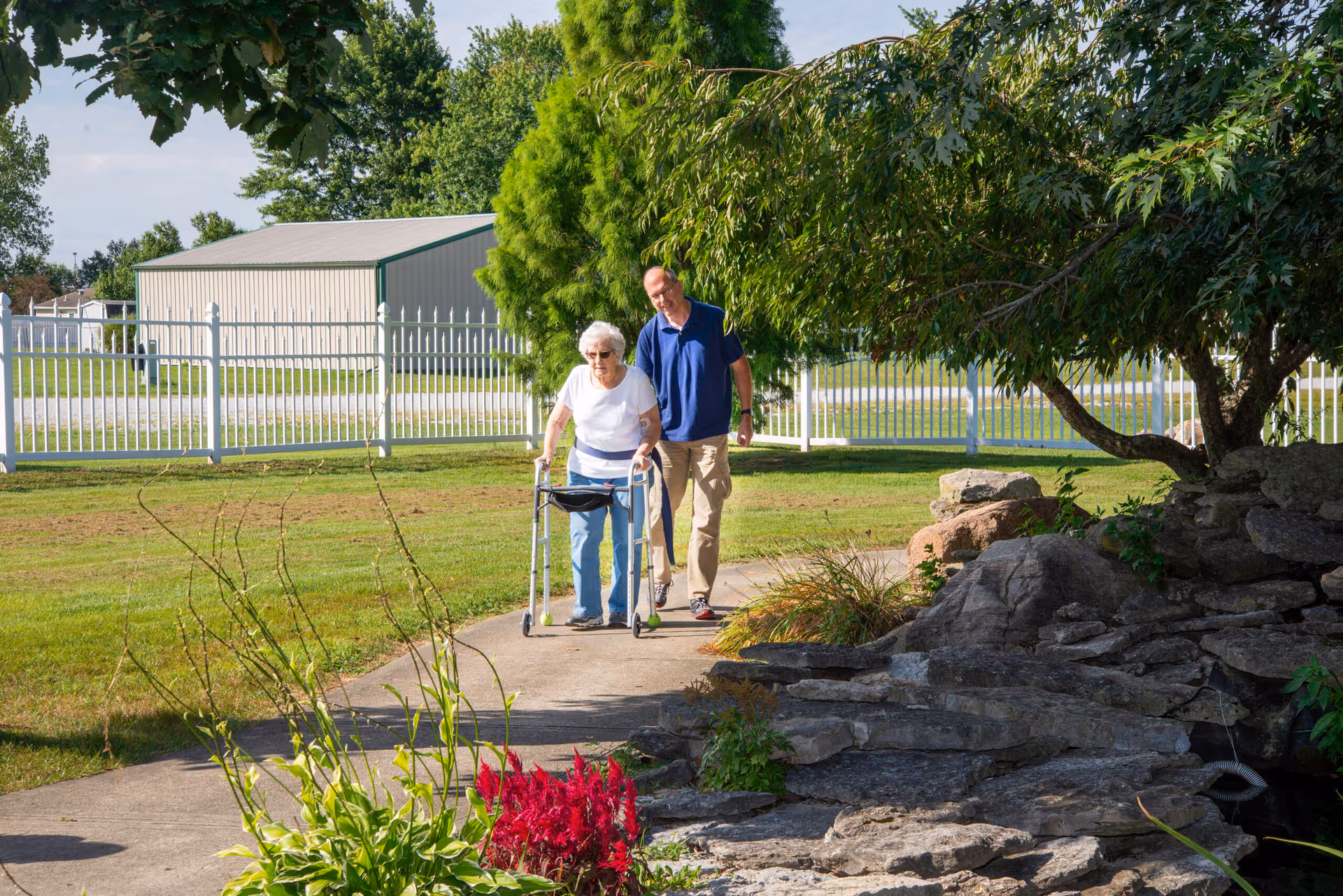 An elderly woman using a walker is accompanied by a man walking beside her on a paved path in a garden area with green grass, trees, and a rock feature. A white fence and a large shed are visible in the background under a clear sky.