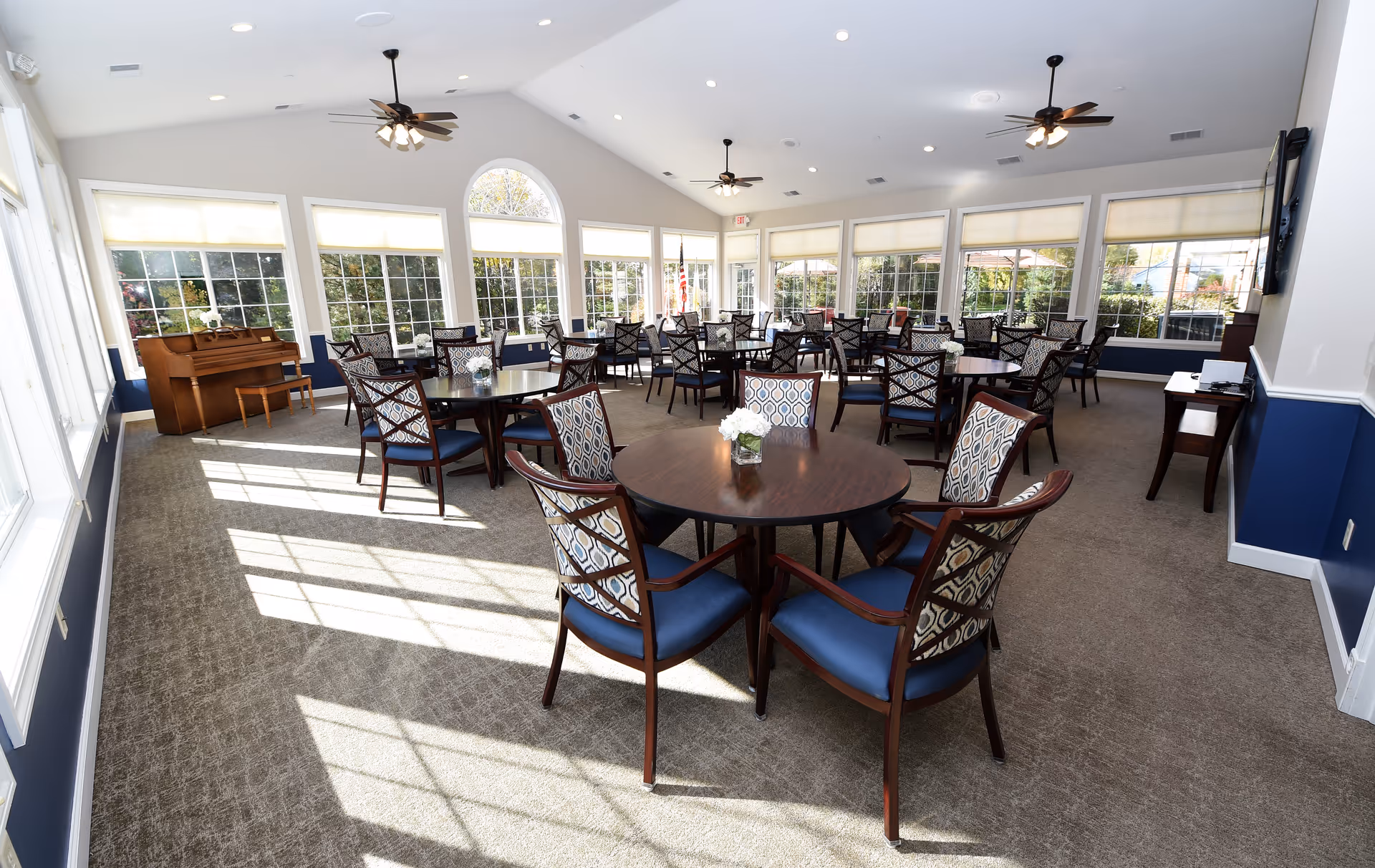 A spacious, well-lit dining room with multiple round wooden tables surrounded by cushioned chairs with patterned backs. Large windows line the walls, allowing natural light to flood the room. Ceiling fans and recessed lighting are installed on the high ceiling. A piano is positioned near the windows on the left side of the room.