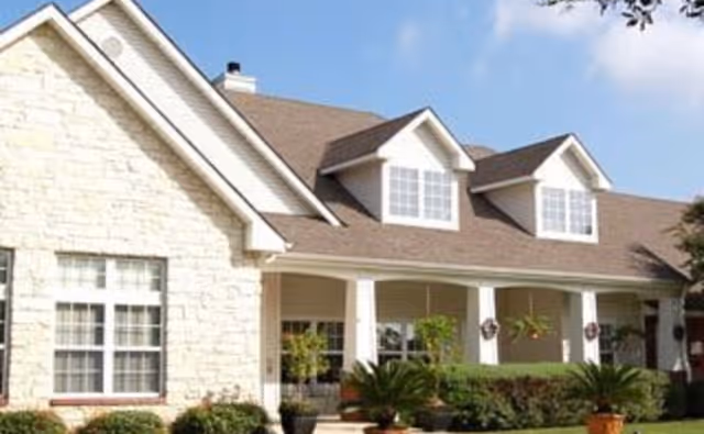Exterior view of a single-story residential-style building with a stone facade and beige siding, featuring multiple windows, a covered porch with white columns, and a well-maintained garden with shrubs and potted plants under a blue sky.
