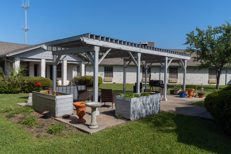 Outdoor patio area at Coastal Palms Nursing & Rehabilitation featuring a wooden pergola with seating underneath, surrounded by planter boxes, potted plants, and green grass. The building with white brick walls and windows is visible in the background under a clear blue sky.