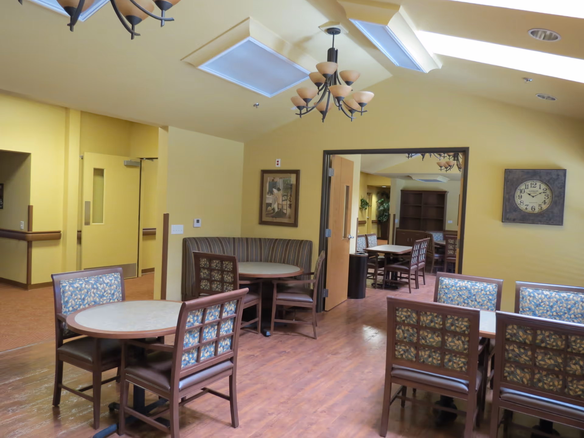 Interior view of a dining area in Amber Grove Place Memory Care with round and rectangular tables surrounded by chairs with patterned upholstery. The walls are painted yellow, and there are chandeliers hanging from the ceiling. A large clock and framed artwork are visible on the walls. An open doorway leads to another room with additional tables and chairs.