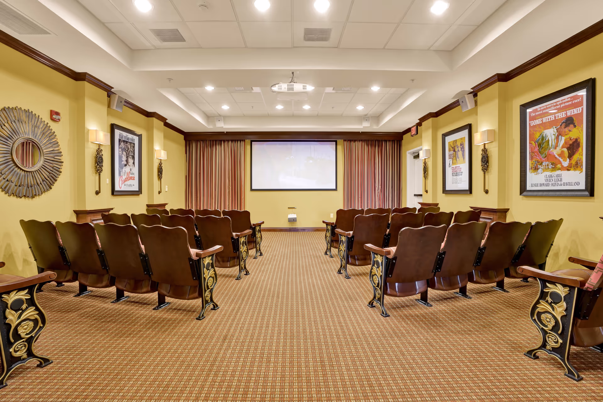 A small theater room with rows of vintage-style wooden seats facing a large projection screen. The room has yellow walls adorned with framed classic movie posters and decorative wall sconces. The carpet is patterned, and the ceiling has recessed lighting and a mounted projector.