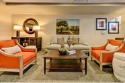 A cozy living room with a beige sofa and two orange armchairs arranged around a wooden coffee table. The coffee table has decorative items including a bowl and books. Behind the sofa, there is a wall with a large painting of a tree and two framed pictures. A round mirror and a lamp are on a side table to the left of the sofa. The room has warm lighting and a carpeted floor.