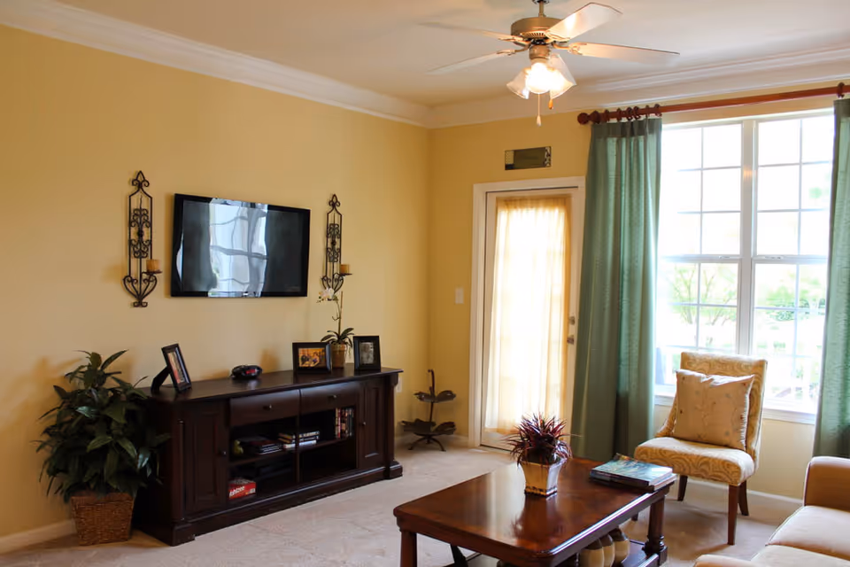 Sunlit living room with a wall-mounted TV above a wooden media console, coffee table, armchair, and large window with green curtains.