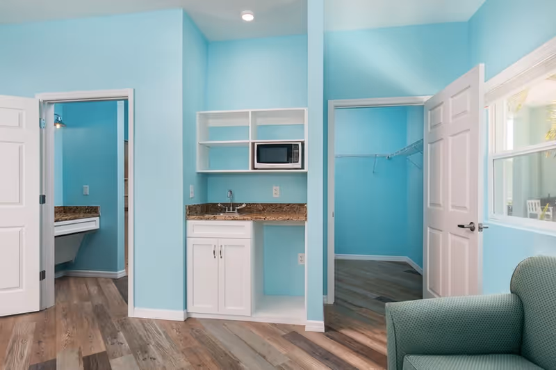 Small studio-style interior with turquoise walls featuring a kitchenette with a microwave and sink, two open doorways and a green armchair.
