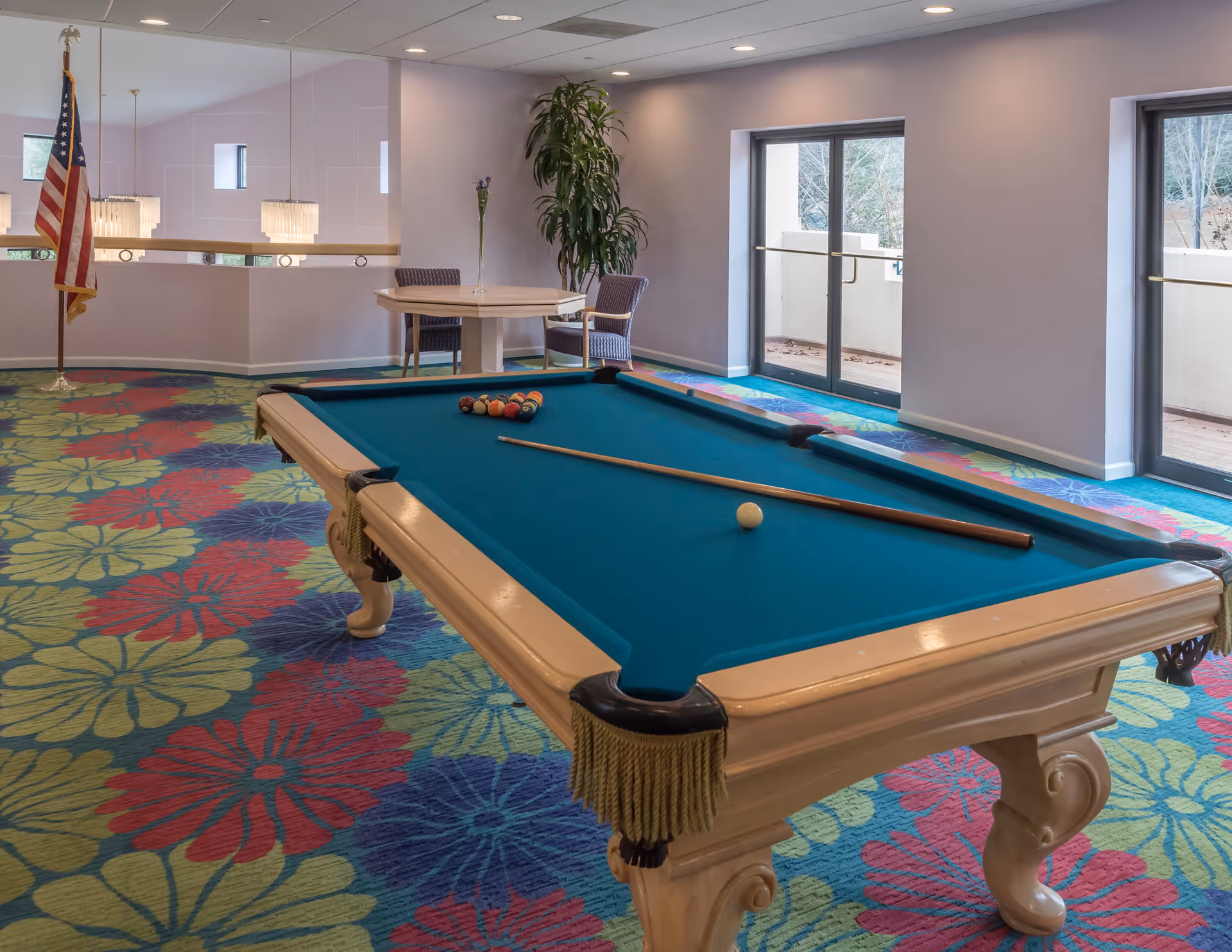 Interior room with a pool table in the center, a cue stick and billiard balls arranged on the table. The room has colorful floral carpet, two chairs and a small round table with a vase and flower, large windows with glass doors leading outside, and an American flag in the corner.